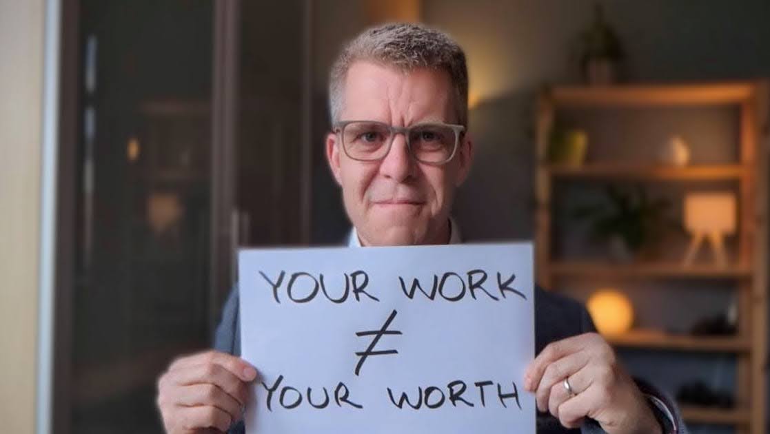 Benjamin at his desk holding a sign saying your work does not equal your worth